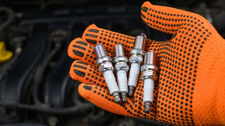 An orange-gloved hand holding four spark plugs in front of a car's engine bay