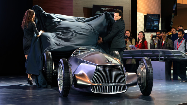 The open-wheeled Infiniti Protoype 9 concept car unveiled at an auto show with a man and woman dressed in black pulling a cover sheet off the car