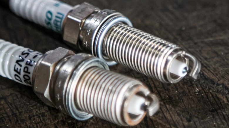 Foreground: Traditional spark plug with shorter thread reach and larger center electrode. Background: Iridium spark plug with longer thread reach and smaller central electrode.