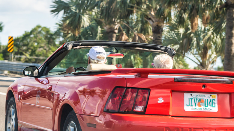 Older couple driving in a red car with Florida license plate.