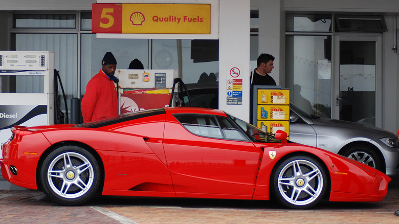 A red Enzo Ferrari fuels up at a Shell gas station while an attendant in a red jacket looks at it