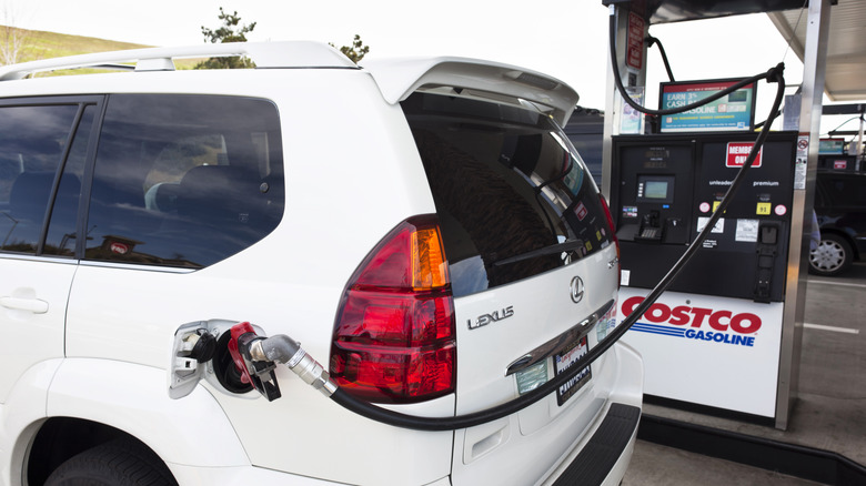 A white Lexus SUV fills up with gas at a Costco, stretching the extra long filler hose around the other side