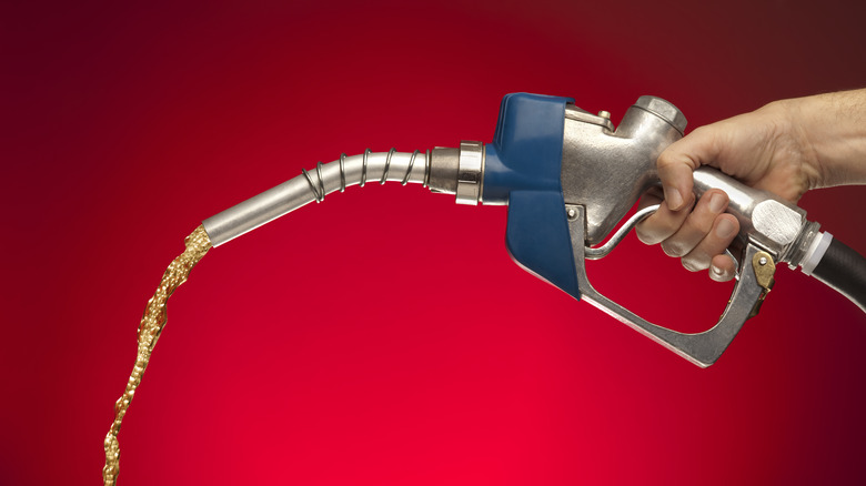 A hand holds a gas pump releasing fuel in midair against a red background