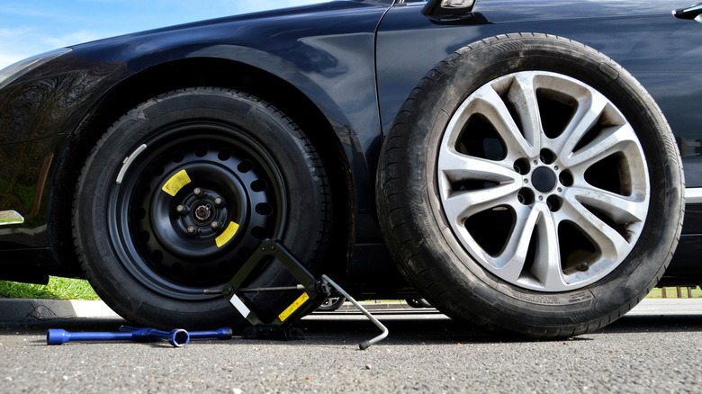 A tire being changed to a space-saver spare tire and aluminum rim following a puncture, with a lug wrench and jack also seen