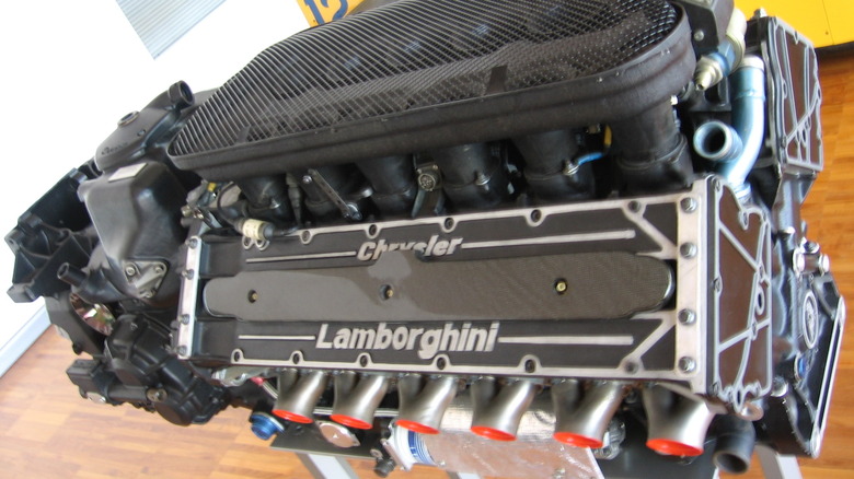 A Chrysler/Lamborghini Formula 1 V12 engine on a stand over a wooden floor, white wall, and yellow cabinets