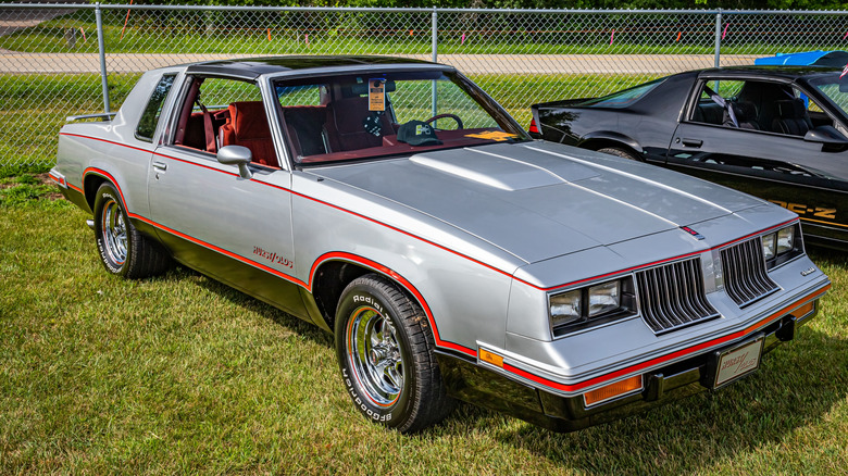 A silver 1983 Hurst Olds Cutlass sitting on green grass in bright sun at a car show.
