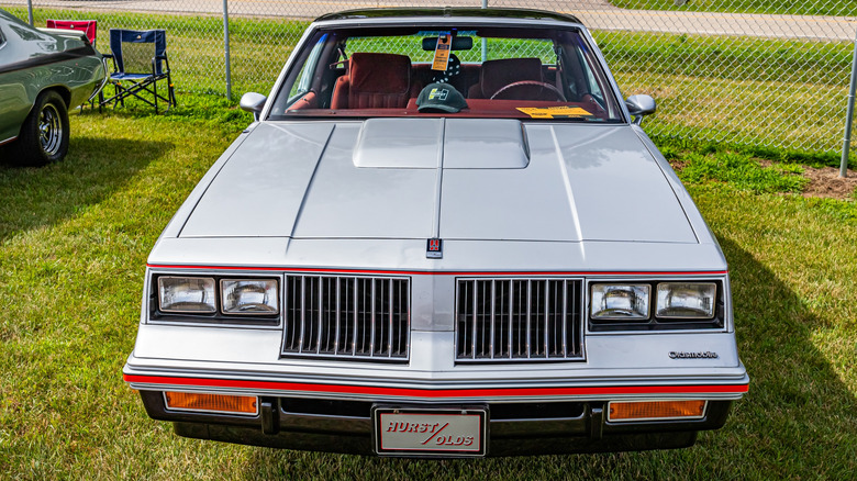 A silver 1983 Hurst Olds Cutlass sits at a car show in sunny lighting.