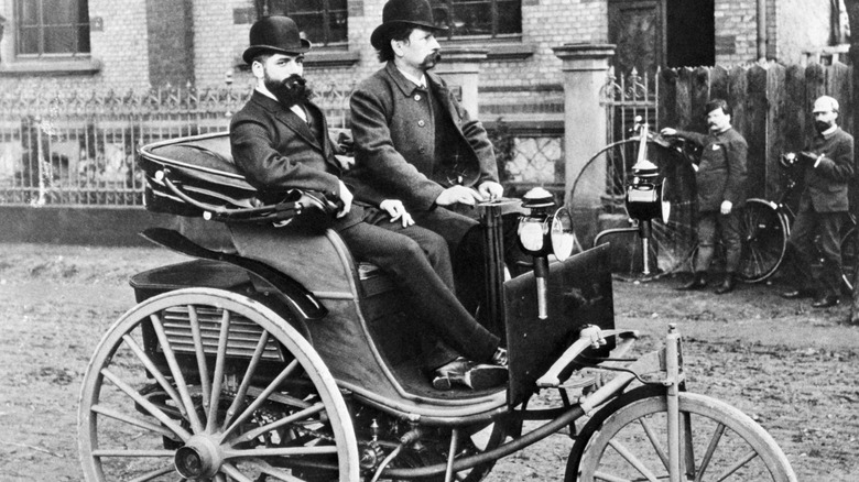 A black and white image of two people in the first Mercedes-Benz, a single-cylinder tricycle, with two more people in the background near bicycles.