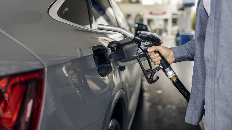 A person prepares to pump gasoline into a grey sedan at a fuel station