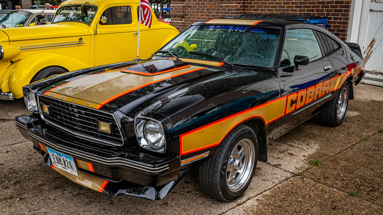 A black and gold 1978 Ford Mustang II Cobra parked next to a yellow hot rod on wet pavement in front of a brick wall at a car show