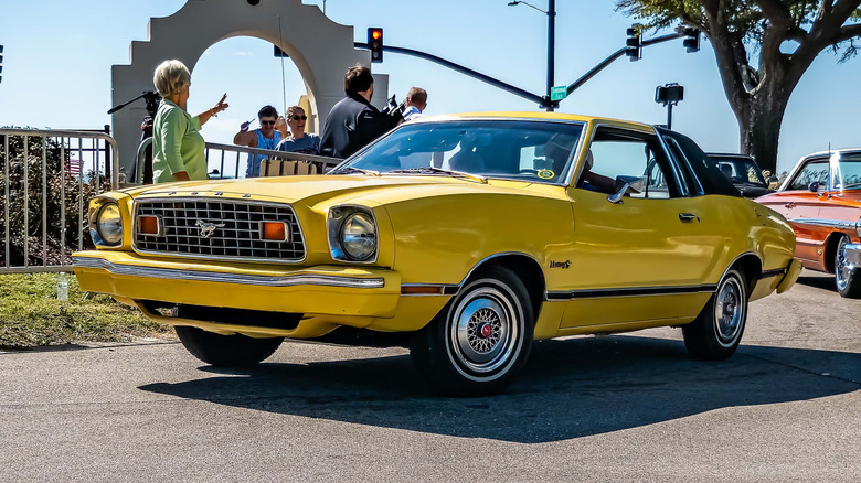 A yellow Ford Mustang II pulling into a car show past a small crowd and a traffic light
