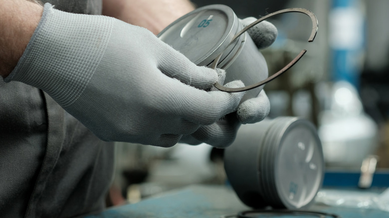 A technician measuring and mounting piston rings to a piston, wearing gray gloves.