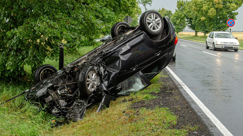 A black SUV overturned on its roof next to a road due to a rollover accident, with a crumpled front and right front door.