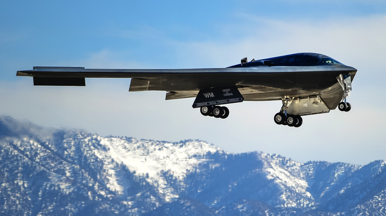 A B-2 Spirit stealth bomber, The Spirit of Hawaii, with its landing gear deployed and snow capped mountains in the background.