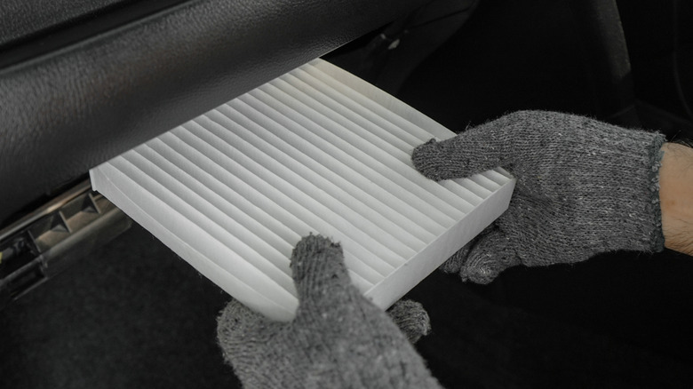 A person's gloved hands inserting a new cabin air filter under the glove box