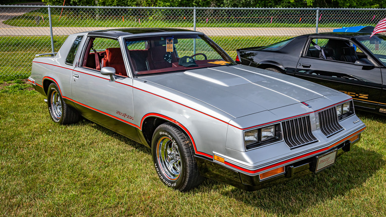 A silver 1983 Hurst Olds Cutlass sitting on green grass in bright sun at a car show.