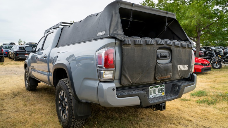 A gray Toyota Tacoma with a fabric bed cap and tailgate protection, parked on grass with other vehicles in the background.