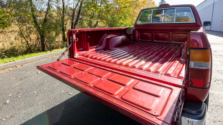 An old red Toyota Tacoma with its tailgate down, parked near a white building, with trees and grass in the background.