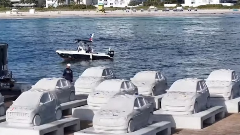 Eight car-shaped concrete bollards drying in the sun on a ship deck, with a beach in the background.