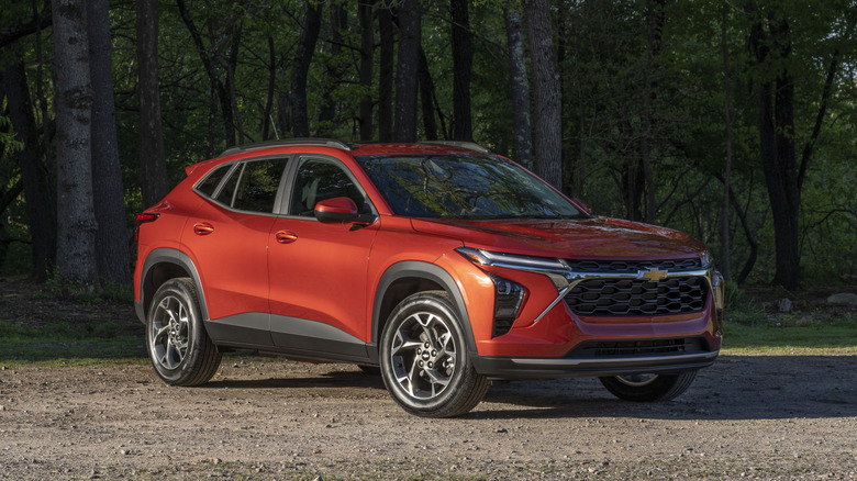 A 2024 Chevrolet Trax LT in Cayenne Orange Metallic parked on a gravel road in front of trees.