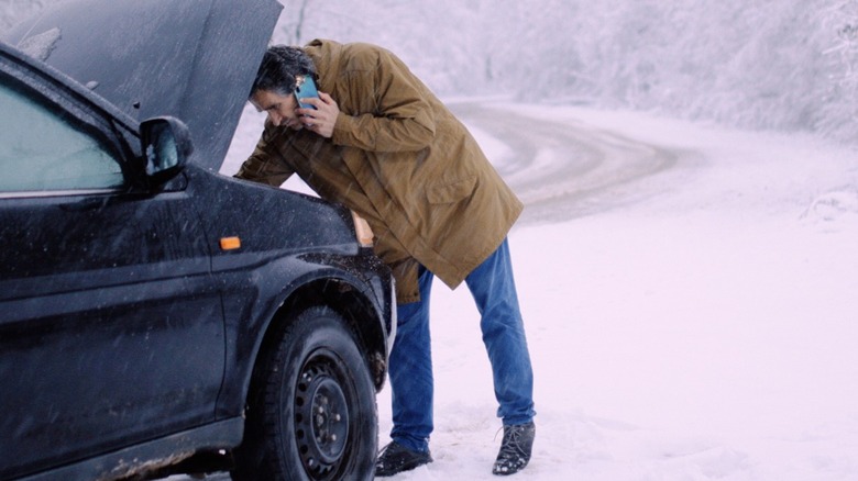 Man talking on phone while looking under hood of car that is broken down along side a snowy road