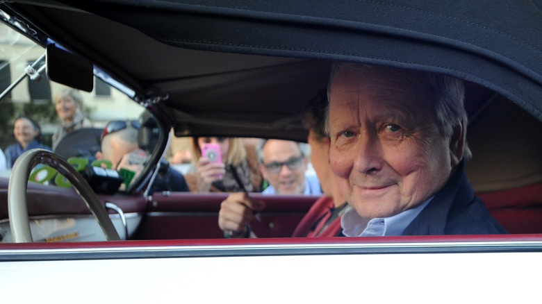 Wolfgang Porsche looks out the driver's side window of a white convertible Porsche 356 with a black top as a crowd on the other side of the car looks and takes pictures