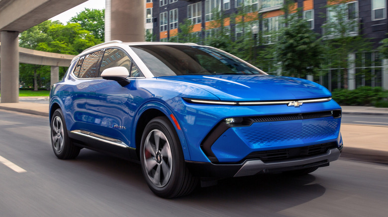 A front 3/4 view of a 2026 Chevrolet Equinox EV LT in Riptide Blue Metallic with a Summit White roof driving in a downtown area