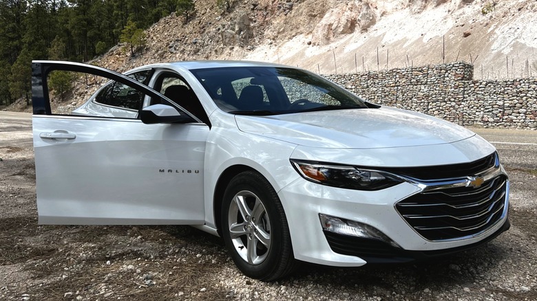 A front-right 3/4 of a white 2020 Chevy Malibu hybrid with its front passenger door open parked next to a road.