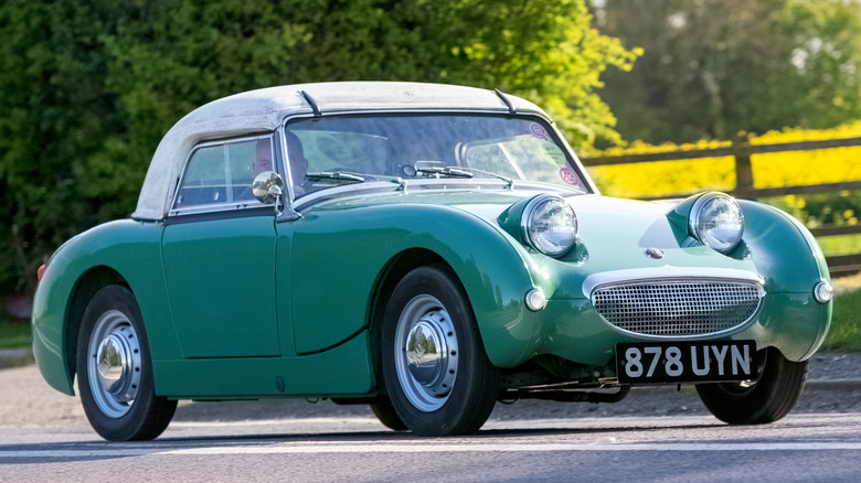A green and white 1960 Austin Healey Sprite on a road with trees, a fence and flowers in the background.