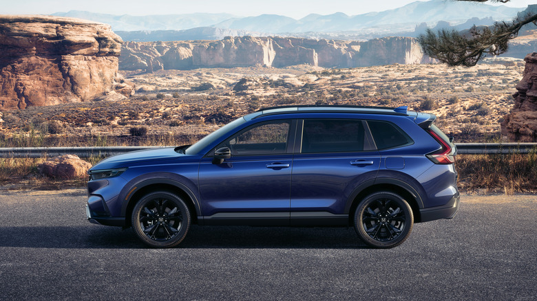 A side profile of a blue 2026 Honda CR-V Hybrid in a parking lot with mountains in the background