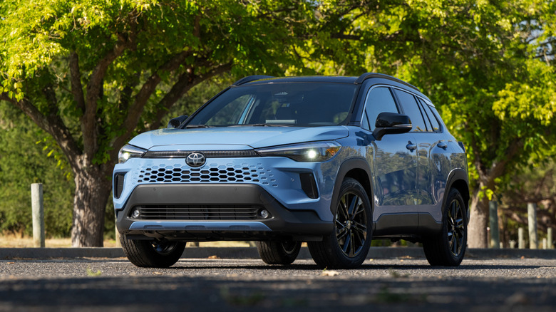 A front left 3/4 of a blue 2026 Toyota Corolla Cross Hybrid in front of trees.