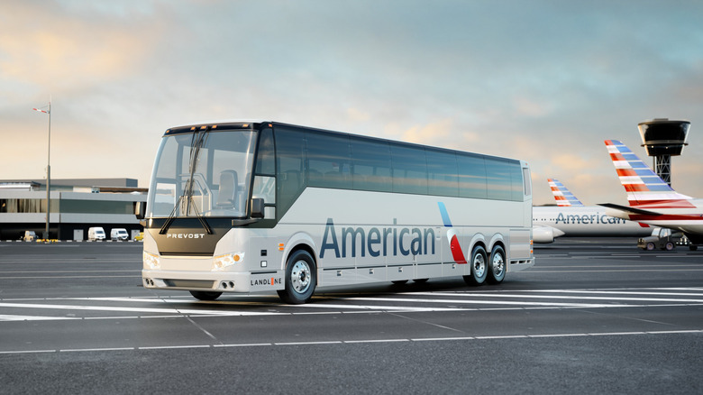 A front three-quarters shot of a white Prevost bus with an American Airlines logo on the side parked on the tarmac in front of an airport terminal with jets