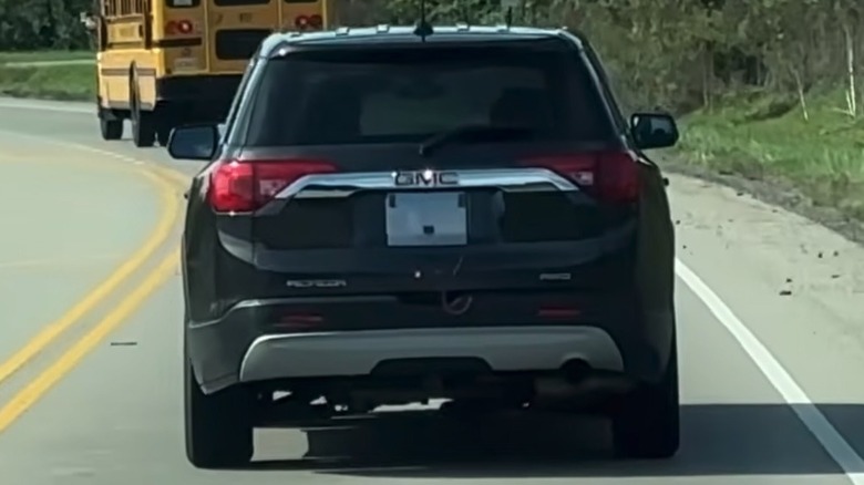 A GMC Acadia drives down the road behind a school bus; the Acadia's license is obscured