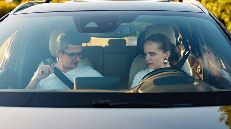A woman driver looking at a male passenger putting on a seatbelt
