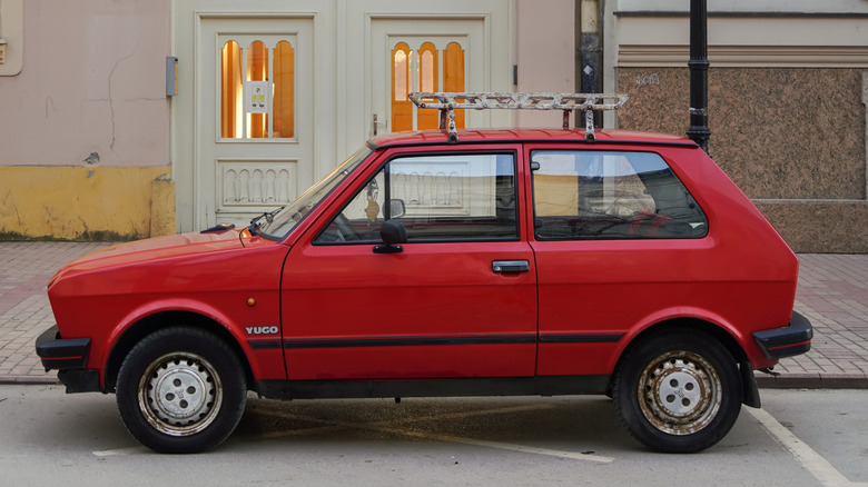 A red Zastava Yugo with a roof rack parked outside a historic building