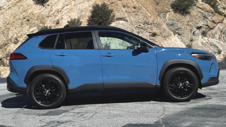 A side view of the blue Corolla Cross Hybrid parked on asphalt in front of a rock mountainside