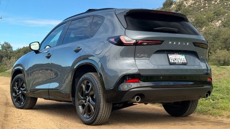 A rear three quarters view of the gray CX-5 parked on a dirt road surrounded by grass in front of a shrubby mountainside