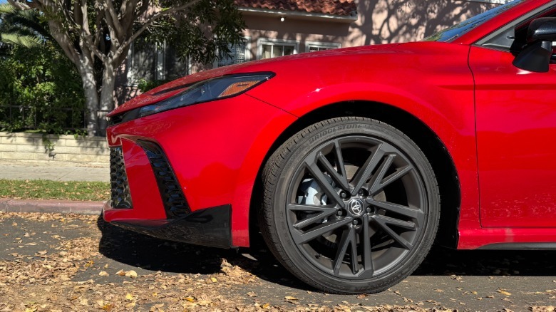 A close-up shot of the front driver's side of the red Camry Nightshade showing the long front overhang parked in front of grass and a tree