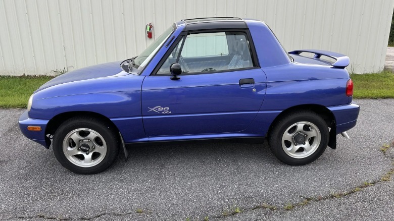 Side view of a blurple Suzuki X-90 parked on asphalt in front of grass and a white wall