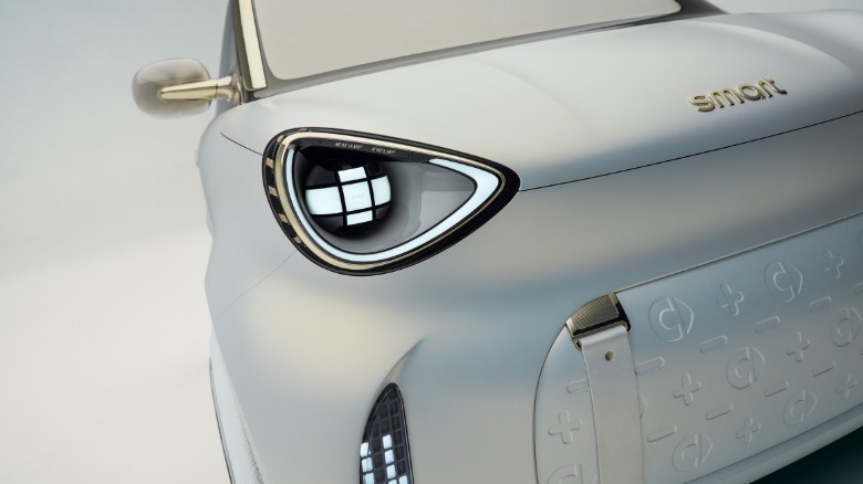 A close-up of the front headlight and 'grille' on the white and gold Smart #2 in a white studio