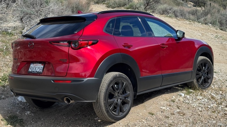 A rear three-quarters shot of the red CX-30 parked on a dirt trail with its rear wheel cocked up in the air in front of shurbs