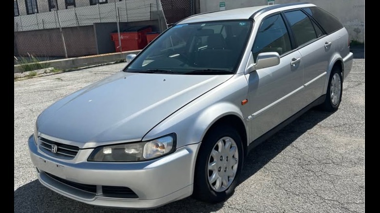 A front three-quarters shot of the silver Accord wagon parked on blacktop in front of a chainlink fence