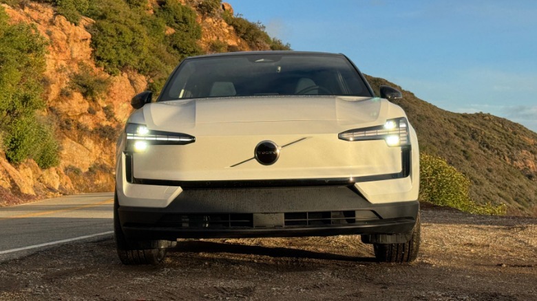 A head-on shot of the white EX30 with its headlights on parked in dirt in front of a rocky shrubby mountainside