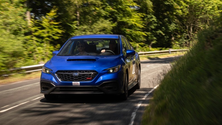 A head-on shot of a blue WRX driving around a corner on a rural road surrounded by trees and grass