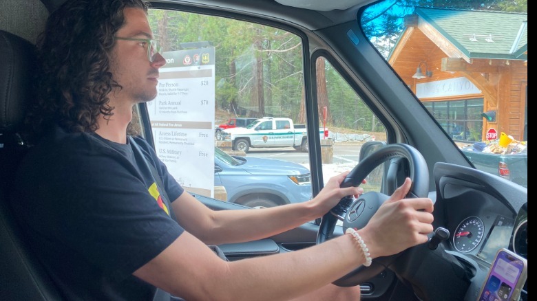 A side view of me driving the van in front of a log cabin and a park ranger truck