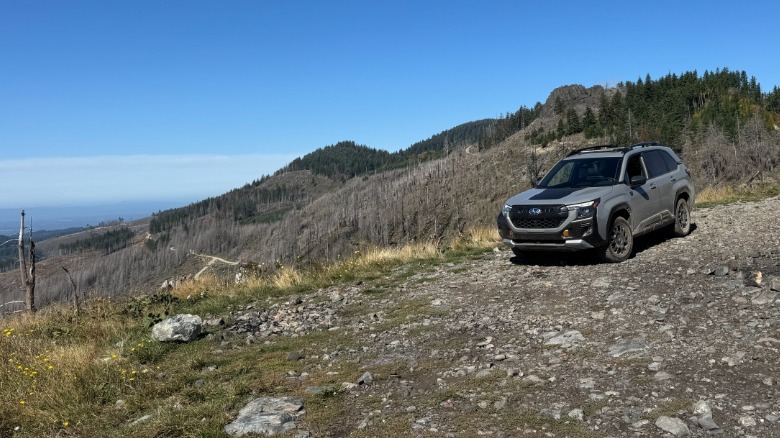 A wide front three quarters shot of the gray Forester Wilderness parked on rocks at the top of a mountain overlooking a view of more mountains