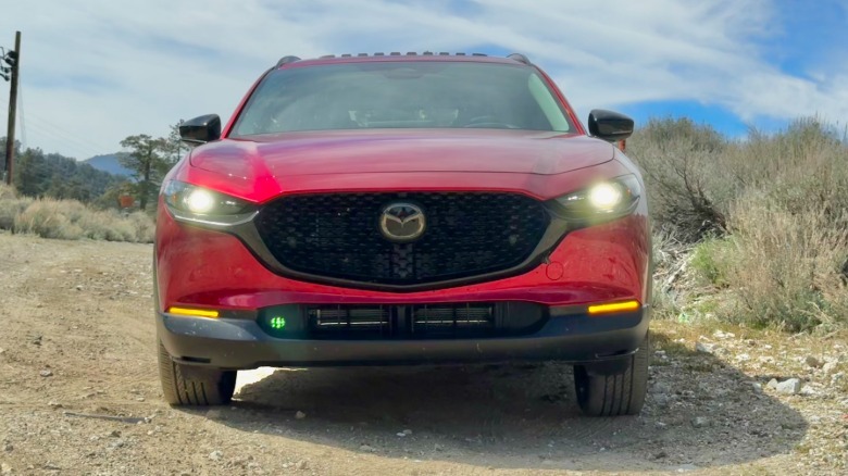 A head-on shot of the read CX-30 with its headlights on while parked on a dirt trail next to shrubs and in front of a partly cloudy sjy