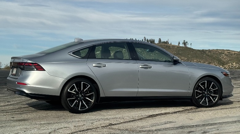 A side view of the silver Accord Hybrid Touring parked on asphalt in front of a cloudy sky and a shrub-covered hill