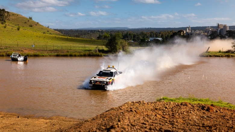 A photo showing the Brataroo three quarters of the way across a body of muddy water approaching a dirt shore