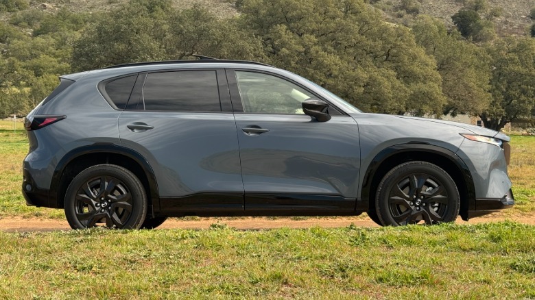 A side view of the gray CX-5 parked on a dirt road surrounded by grass in front of a shrubby mountainside